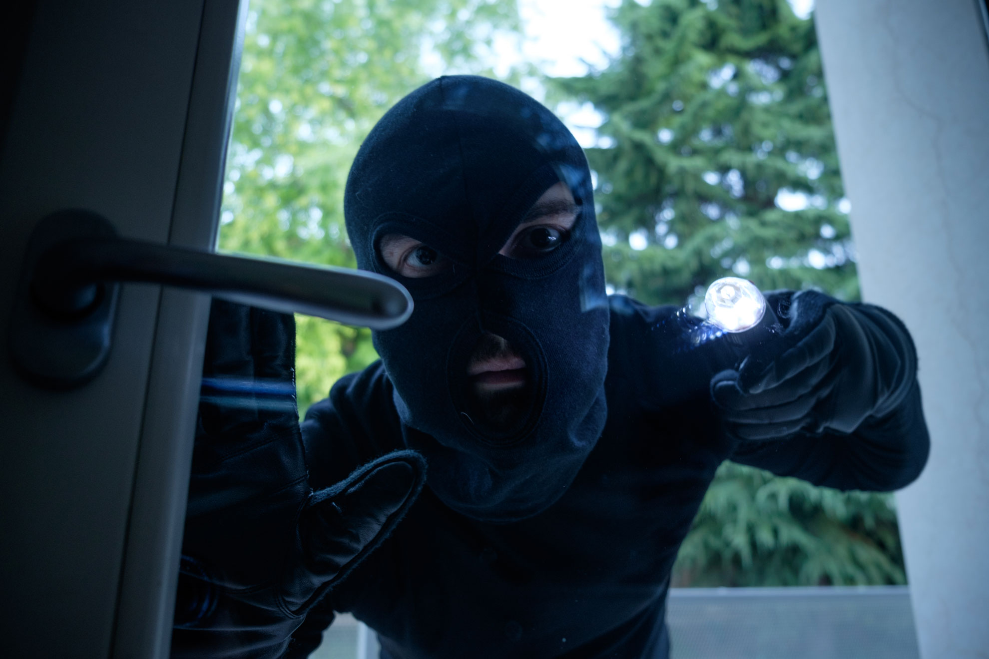 Einbrecher mit Sturmmaske und Taschenlampe an einer Glastür